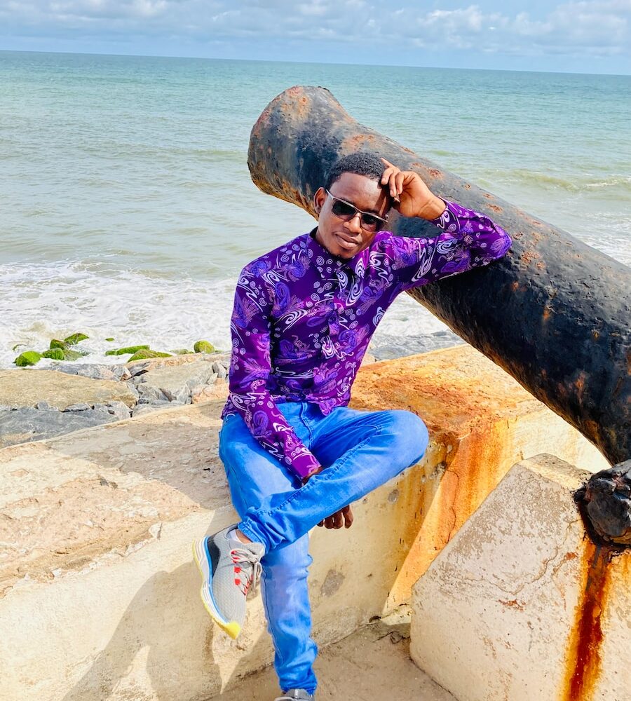 a man in a purple shirt and blue pants sitting on a rock near the ocean